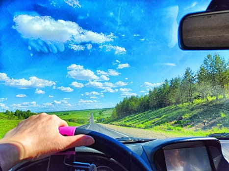 Car salon, windshield, hand of woman on steering wheel and landscape. View from seat of driver on nature with Road, tree and blue sky at sunny day