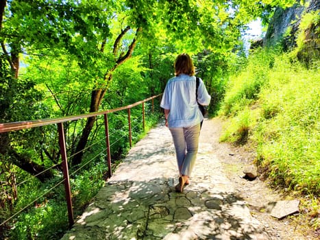 A woman strolls peacefully down a stone pathway enveloped by vibrant green foliage on a sunny day.