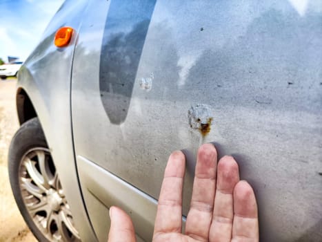 A person examines rust and scratches on silver car parked in a bright outdoor setting, focusing on the damaged area