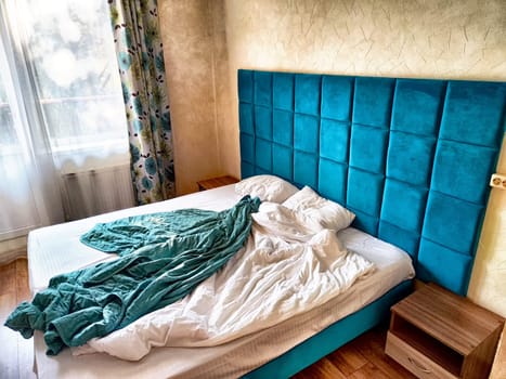 A cozy bedroom displays an unmade bed with crumpled blankets and a bright blue headboard, illuminated by soft morning light.