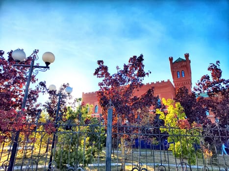 Colorful autumn foliage surrounds a historic red castle, casting beautiful shadows in the tranquil park on a sunny day.