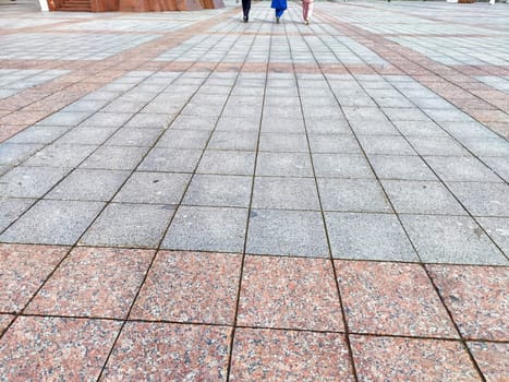 Two individuals walk across a large stone pavement in an open plaza, enjoying a sunny day in the city environment.