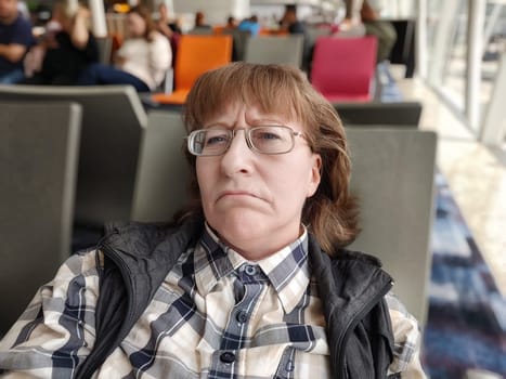A tired woman, wearing glasses and casual attire, sits in an airport terminal surrounded by other travelers, showing signs of impatience as she waits for her flight.