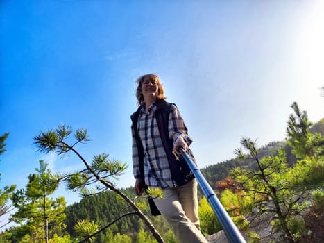 A middle-aged female tourist blogger takes a selfie on a travel. A woman takes a selfie with a selfie stick in forest and blue sky on background