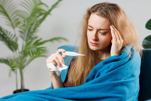 a sick girl with a cold or flu wrapped in a blue blanket measures her temperature at home, symptoms, treatment of viral diseases