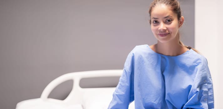 Young female patient in hospital ward,dressed in blue gown,sitting on bed and waiting to be examined by doctor,smiling softly and satisfied with service and services provided,individual hospital room