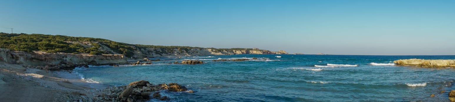 Gentle waves lap against the rocky coast, under a clear blue sky, creating a peaceful seaside escape.
