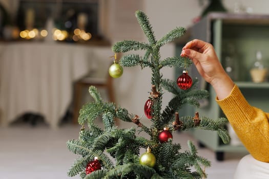 Woman in yellow sweater decorating small Christmas tree with toys in cozy home atmosphere. merry christmas