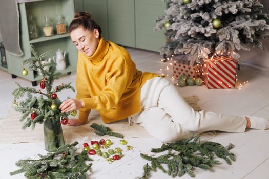 Woman in yellow sweater decorating small Christmas tree with toys in cozy home atmosphere. merry christmas