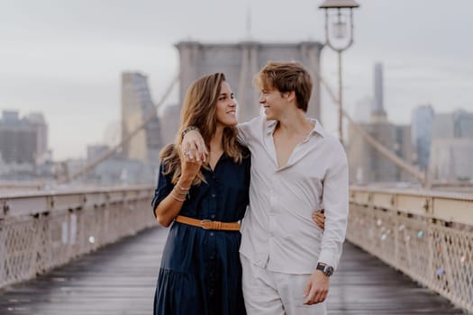 A couple strolls hand in hand on Brooklyn Bridge, surrounded by a stunning skyline and beautiful morning light
