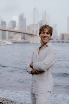 A cheerful young man strikes a pose by the shimmering water, with a breathtaking city skyline in the background. Brooklyn bridge, Manhattan in New York.
