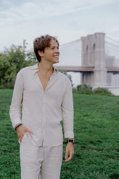 A stylish individual stands confidently in a vibrant green park with the Brooklyn Bridge in the background