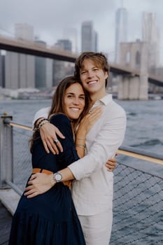 A joyful couple embraces by the waterfront near Brooklyn Bridge, with NYC skyline in the background.