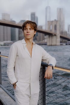 A fashionable young man in a white outfit poses by the Brooklyn Bridge, capturing urban sophistication.