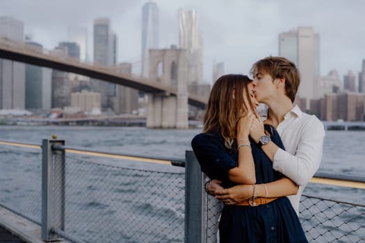 A joyful couple embraces by the waterfront near Brooklyn Bridge, with NYC skyline in the background.