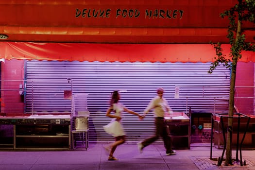 In a lively nighttime setting, a couple joyfully races past a closed food market, full of excitement and fun