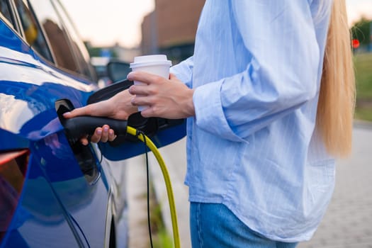 Woman is connecting a charging cable to her electric vehicle in a parking area, holding a cup of coffee.
