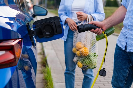 An electric car is parked and charged by man, while a woman carries groceries nearby