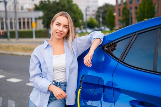 Blonde woman smiles while relaxing beside her electric vehicle as it charges in the street
