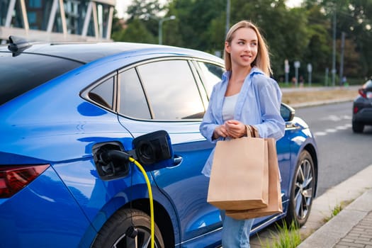 Woman parks her electric car and begins charging it while carrying shopping bags in the evening