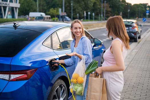 Two women chatting and holding bags near an electric car charging in a parking area during daylight hours