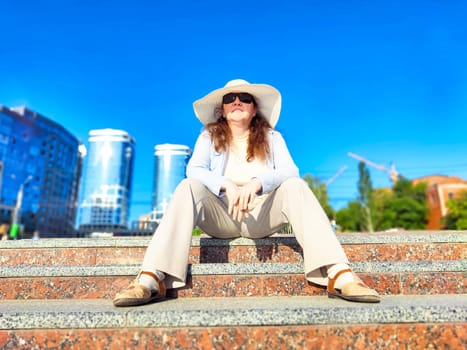 A woman sits comfortably on stone stairs in city, enjoying the warmth of a sunny day while wearing a wide-brimmed hat
