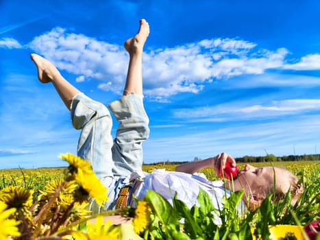 A carefree child reclines in a blooming dandelion field under a bright blue sky, playfully munching on a red apple.