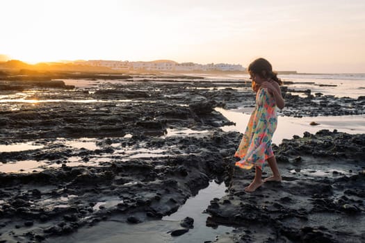 little girl in summer dress walking barefoot at sunset by the volcanic rocks of the beach of Famara in Lanzarote, Canary Islands, Spain, concept of travel and wild nature
