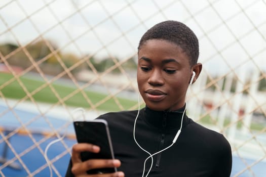 young focused ethnic female text messaging on cellphone while listening to music with earphones against grid fence in city
