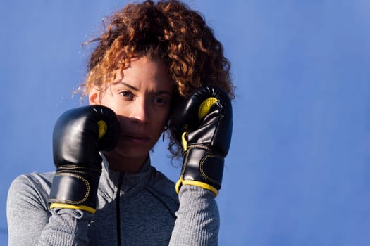 determined adult female boxer with curly hair ready to fight on boxing gloves during workout on blue background, concept of strength and healthy lifestyle, copyspace for text