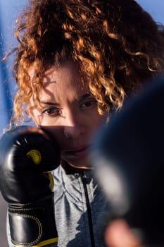 close up portrait of a determined adult female boxer with curly hair ready to fight on boxing gloves during workout on blue background, concept of strength and healthy lifestyle