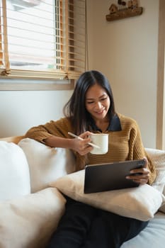 Charming young woman holding cup of hot beverage using digital tablet on sofa.