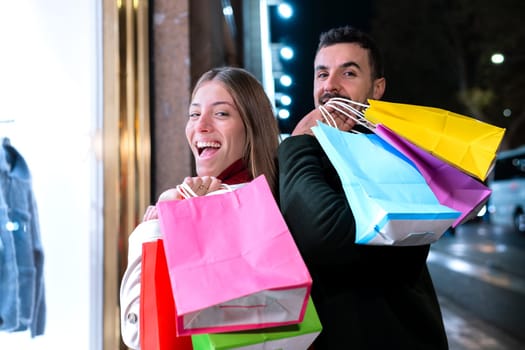 Smiling young couple holding colorful shopping bags, enjoying a night out in the city. They radiate excitement and joy, perfect for concepts related to shopping, lifestyle, and urban experiences