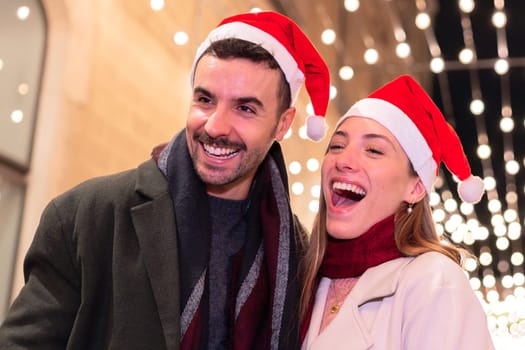 Cheerful couple wearing Santa hats, laughing and enjoying festive holiday lights on a winter night. Perfect for Christmas, holiday season, celebration, and joyful moments in a city setting.