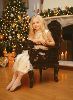 Happy little girl child in dress sitting on a armchair near Christmas tree and fireplace with holiday lights at home
