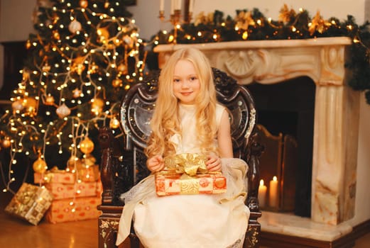 Happy little girl child in dress with festive gift box sitting near Christmas tree and fireplace with holiday lights at home