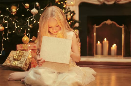 Happy little girl child opens festive gift box sitting near Christmas tree and fireplace with holiday lights at home