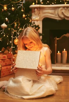 Happy little girl child opens festive gift box sitting near Christmas tree and fireplace with holiday lights at home