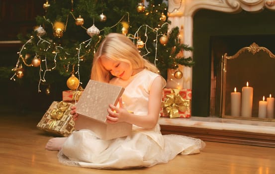 Happy little girl child opens festive gift box sitting near Christmas tree and fireplace with holiday lights at home