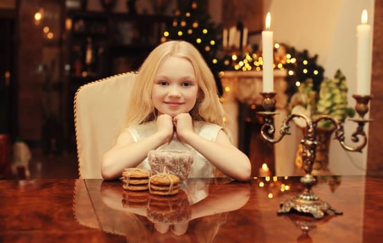Christmas night, happy little girl kid makes a wish sitting at the table with sweets and burning candles at home, garland lights background