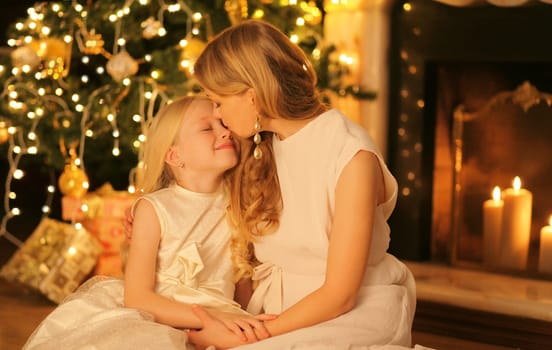 Happy mother kissing her smiling child daughter near Christmas tree and fireplace at home