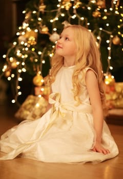 Happy little girl child in dress looking up makes a wish with near Christmas tree with holiday lights at home