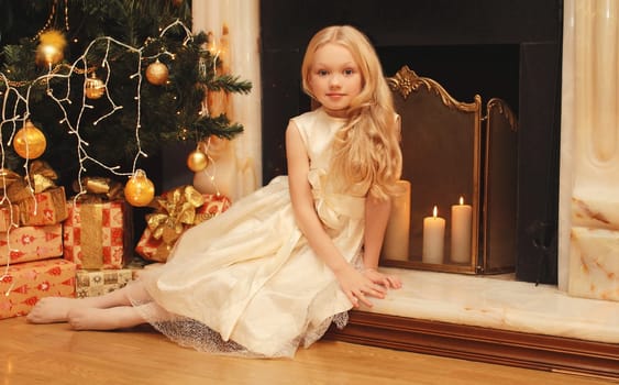 Happy little girl child in dress with festive gift box sitting near Christmas tree and fireplace with holiday lights at home