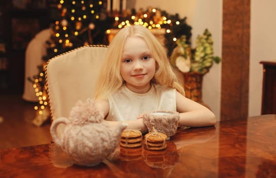 Christmas night, happy little girl kid makes a wish sitting at the table with sweets at home, garland lights background