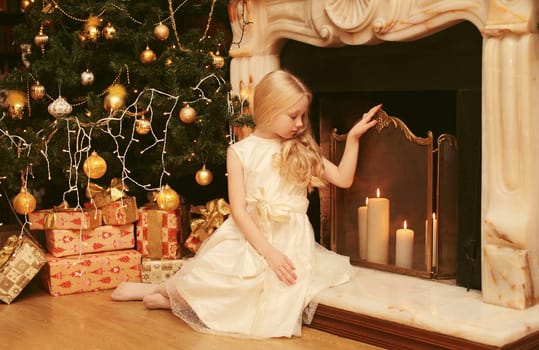 Happy little girl child in dress with festive gift box sitting near Christmas tree and fireplace with holiday lights at home