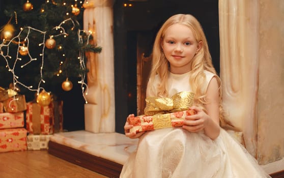 Happy little girl child in dress with festive gift box sitting near Christmas tree and fireplace with holiday lights at home