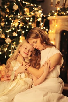 Happy young mother hugs child daughter near Christmas tree with lights at home