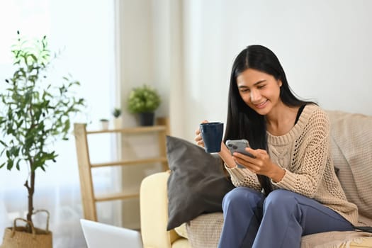 Pretty young woman sitting on couch holding a coffee cup and reading messages on smartphone.