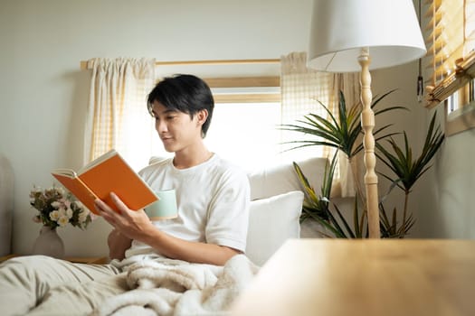 Peaceful young adult man drinking hot coffee and reading book in living room.