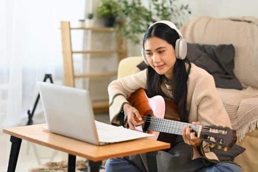 Focused young woman playing guitar while watching online tutorial on laptop.